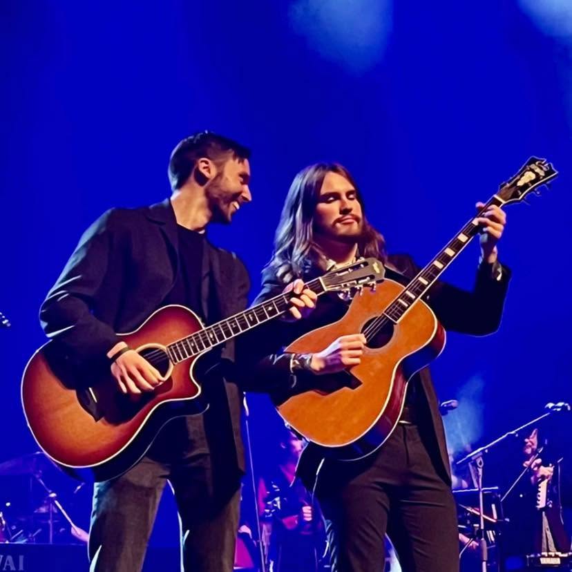 The Murphy brothers on stage in matching dark suits, both playing acoustic guitars under blue stage light.