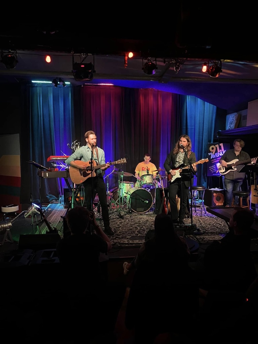 The brothers performing with a full band on a club stage: acoustic guitar and keys at left, drums centre, electric guitar and bass at right.