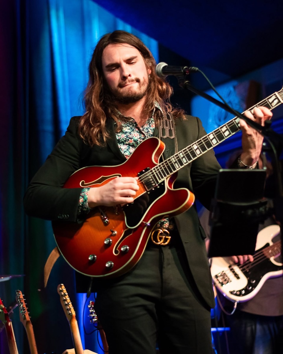 One of the brothers playing a semi-hollow electric guitar at the microphone under blue stage light.